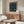 May include: William Morris exhibition poster featuring intricate dark blue pattern with birds and foliage in a light wood frame, displayed above a mid-century walnut credenza in a minimalist living room with cream walls, white sculptural chair, glass coffee table, and modern brass floor lamp.