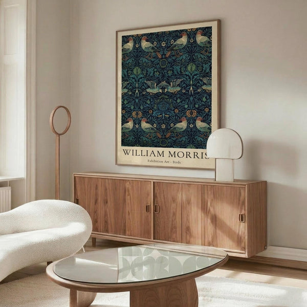 May include: William Morris exhibition poster featuring intricate dark blue pattern with birds and foliage in a light wood frame, displayed above a mid-century walnut credenza in a minimalist living room with cream walls, white sculptural chair, glass coffee table, and modern brass floor lamp.