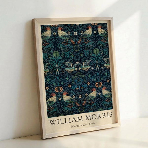 May include: Framed art print featuring William Morris's decorative pattern with birds and botanical elements in deep blue, teal, and green tones. The design shows detailed birds with orange-red heads among intricate flowering vines and foliage. Text reads "WILLIAM MORRIS Exhibition Art - Birds" below the artwork. Natural wood frame displayed against white wall, leaning on surface with soft lighting creating gentle shadows.