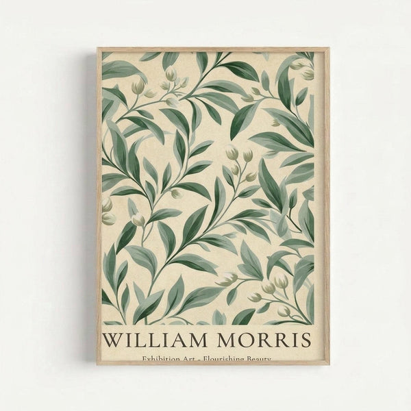 Framed botanical poster with green leaves and buds on cream background in natural wood frame, displayed against white wall. The design features flowing branches with oval leaves in various shades of green, creating an organic pattern. Text at bottom reads "WILLIAM MORRIS" with subtitle "Exhibition Art - Flourishing Beauty" in dark lettering.