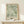 May include: Framed botanical poster with sage green background featuring flowing white and green leaf patterns in William Morris style, displayed on light wood console table beside small wrapped gift with twine and evergreen sprig against cream wall