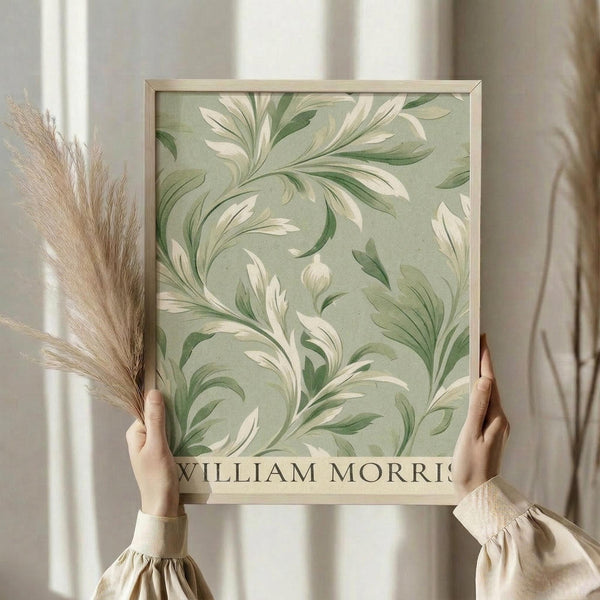 May include: Person in cream sweater holding framed botanical print with flowing green and white leaf patterns against sage green background, standing near window with dried pampas grass, natural lighting creating soft shadows on neutral curtains