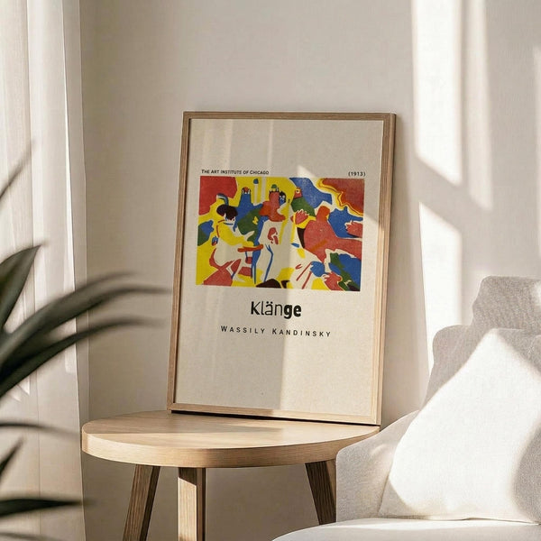 May include: Framed art print featuring Wassily Kandinsky's abstract artwork with vibrant red, yellow, blue, and green shapes depicting stylized figures in motion. The poster shows museum exhibition text reading "Klänge" and "The Art Institute of Chicago (1913)". Light wood frame sits on round wooden side table next to beige upholstered chair, with plant leaves visible on left side in bright, naturally lit room with white walls.
