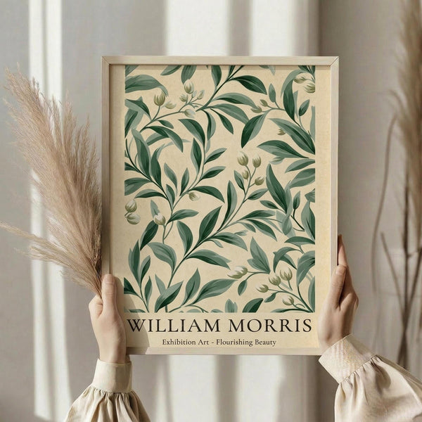 May include: Hands holding a framed botanical art print featuring green leafy branches with small buds on a cream background, displayed against white curtains with dried pampas grass visible to the left, person wearing a beige ruffled sleeve top