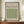 May include: Framed poster with beige background displaying green geometric diaper pattern in grid layout, attributed to William Morris and Smithsonian Institution, dated 1870. Wood frame sits on wooden sideboard next to white ceramic vase and stacked books against cream wainscoted wall.