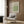 May include: Framed William Morris poster with green geometric diaper pattern design hanging on wood-paneled wall above cream upholstered armchair with patterned pillow. Modern brass floor lamp and dark side table with open book visible in neutral-toned room setting with curtained window.