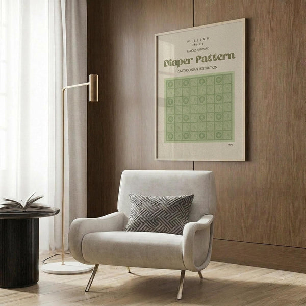 May include: Framed William Morris poster with green geometric diaper pattern design hanging on wood-paneled wall above cream upholstered armchair with patterned pillow. Modern brass floor lamp and dark side table with open book visible in neutral-toned room setting with curtained window.
