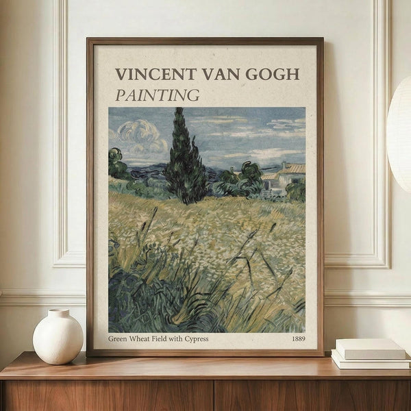 May include: Framed Van Gogh reproduction poster showing a golden wheat field with tall dark cypress tree and cloudy sky, displayed on wooden sideboard next to white ceramic vase and stacked books in room with cream walls and white trim molding.