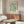 May include: Framed botanical art print with flowing green and cream leaves on sage background, displayed above mid-century walnut credenza in minimalist living room. White sculptural chair, glass oval coffee table, and modern brass floor lamp complete the neutral-toned interior scene.