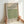May include: Hands holding a framed art print featuring a green geometric pattern with repeating squares and circular motifs. The print has cream background with text reading "WILLIAM Morris" and "FAMOUS ARTWORK Diaper Pattern SMITHSONIAN INSTITUTION" and shows the year 1870. Natural wood frame against white wall in soft lighting.