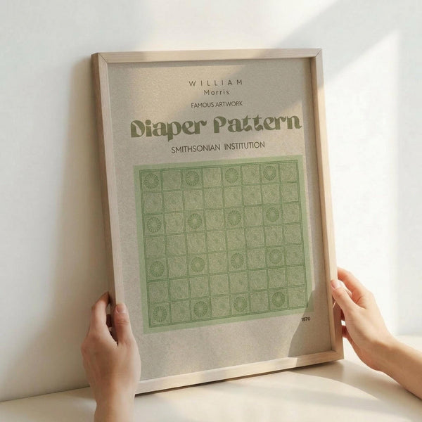 May include: Hands holding a framed art print featuring a green geometric pattern with repeating squares and circular motifs. The print has cream background with text reading "WILLIAM Morris" and "FAMOUS ARTWORK Diaper Pattern SMITHSONIAN INSTITUTION" and shows the year 1870. Natural wood frame against white wall in soft lighting.