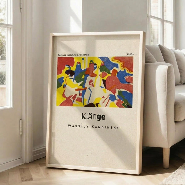 May include: Framed art print leaning against white wall next to beige upholstered armchair in bright living room. Print features colorful abstract artwork with bold red, yellow, blue, and green shapes depicting stylized figures. Museum-style poster format with cream matting, thin light wood frame. Natural sunlight streaming through tall windows creates soft shadows on hardwood floor.