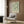 May include: Framed botanical poster with green foliage pattern on cream background mounted on wood-paneled wall. Modern beige upholstered armchair with geometric throw pillow sits below artwork. Brass floor lamp with white marble base stands nearby next to dark round side table with open book. Natural lighting from window with sheer curtains illuminates warm-toned contemporary room setting.