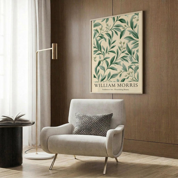 May include: Framed botanical poster with green foliage pattern on cream background mounted on wood-paneled wall. Modern beige upholstered armchair with geometric throw pillow sits below artwork. Brass floor lamp with white marble base stands nearby next to dark round side table with open book. Natural lighting from window with sheer curtains illuminates warm-toned contemporary room setting.