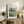 May include: Framed Van Gogh reproduction poster displayed on rustic wooden sideboard in neutral-toned living room. Print shows wheat field landscape with prominent dark cypress tree against cloudy sky in post-impressionist style. Styled with fern plant, cream cushioned chair, ceramic vase, and stacked books creating cozy home decor scene.