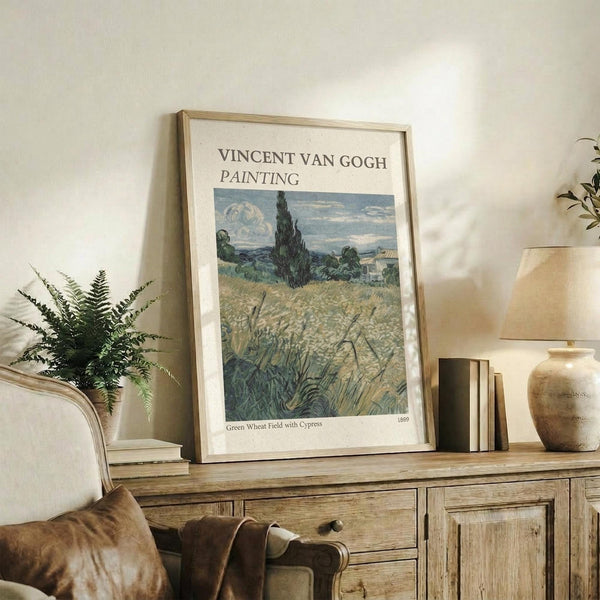 May include: Framed Van Gogh reproduction poster displayed on rustic wooden sideboard in neutral-toned living room. Print shows wheat field landscape with prominent dark cypress tree against cloudy sky in post-impressionist style. Styled with fern plant, cream cushioned chair, ceramic vase, and stacked books creating cozy home decor scene.