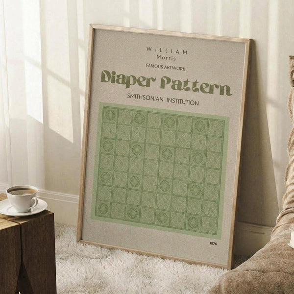 May include: Framed art print with green geometric diaper pattern design in light wooden frame, leaning against white wall corner. Print shows William Morris artwork with repeating circular motifs in squares on cream background, dated 1870. Scene includes wooden side table with white coffee cup and saucer, beige textured throw blanket, and white shag rug. Soft natural lighting creates window shadows on wall.