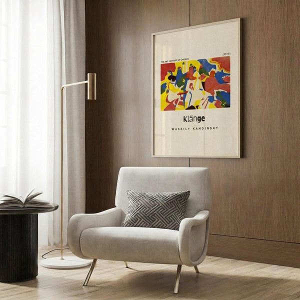 May include: Framed art poster with colorful abstract design featuring bold red, blue, and yellow shapes displayed on wood paneled wall above beige upholstered armchair with geometric patterned pillow. Modern brass floor lamp and dark round side table with open book visible in contemporary neutral-toned room setting.