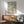 May include: Framed botanical art print with green leafy pattern hanging on white wall above wooden credenza with table lamp. Mid-century modern sage green upholstered armchair with wooden frame sits nearby. Warm sunlight creates shadows across the minimalist living room scene with neutral tones and natural wood furniture.