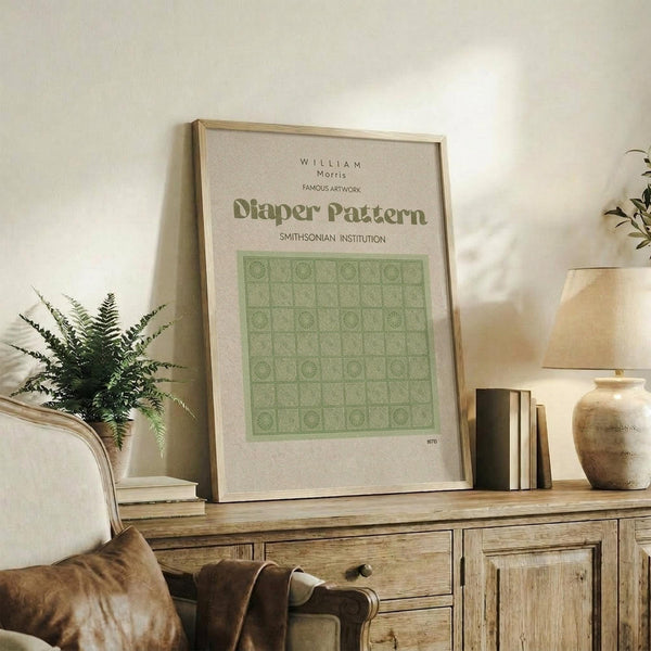 May include: Framed poster with green geometric pattern grid displayed on rustic wooden sideboard alongside fern plant, stacked books, and ceramic vases in warm, naturally-lit living room setting with beige upholstered chair and neutral wall backdrop.