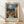 May include: Framed poster print featuring a Mediterranean village scene in Van Gogh's distinctive swirling style, with warm yellow and orange buildings topped with terracotta roofs beneath a dramatic blue night sky with circular, spiraling clouds around a bright moon. The artwork shows cobblestone streets winding between rustic houses and a tall minaret or tower. Natural wood frame with cream matting displays the poster against white walls on light wooden flooring with soft natural lighting.