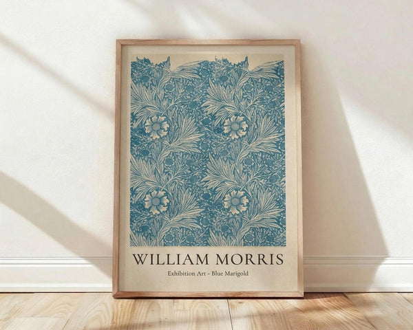 May include: A framed art print featuring an intricate blue and cream botanical pattern with marigold flowers, ferns, and foliage in William Morris's distinctive Arts and Crafts style. The print is displayed in a light wood frame, leaning against a white wall on natural wood flooring with soft natural lighting creating gentle shadows.