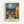 May include: Framed poster displaying a Van Gogh-inspired painting of a Mediterranean village at night with swirling sky, bright moon, terra cotta roofed buildings, and church bell tower. Features warm yellows and oranges contrasting with deep blues, mounted on white wall in light wood frame with French text below artwork.