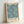 May include: Framed poster with blue and cream botanical pattern featuring marigold flowers and flowing leaves in William Morris Arts and Crafts style. Light wood frame sits propped against white wall, showing natural wood grain and museum-style text label at bottom. Soft natural lighting creates gentle shadows.
