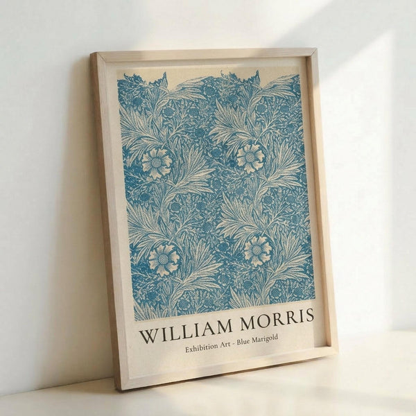 May include: Framed poster with blue and cream botanical pattern featuring marigold flowers and flowing leaves in William Morris Arts and Crafts style. Light wood frame sits propped against white wall, showing natural wood grain and museum-style text label at bottom. Soft natural lighting creates gentle shadows.