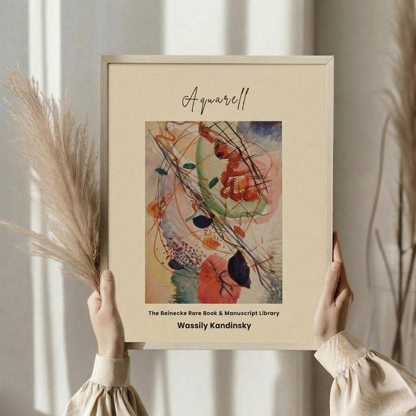 May include: Person in cream sweater holding framed abstract watercolor artwork featuring vibrant swirls of red, orange, green, and blue paint with dynamic black brushstrokes. Frame has light wood finish with cream matting. Dried pampas grass visible on left side against soft natural lighting from window.