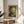 May include: Van Gogh self-portrait print in thin gold frame displayed on round wooden side table next to beige upholstered chair. Artwork shows the artist with reddish beard and hair against swirling blue-green background in his distinctive Post-Impressionist style. Soft natural light creates shadows across white wall. Green plant leaves visible in corner.