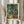 May include: Person in cream ruffled sleeves holding framed Van Gogh poster featuring swirling green and blue cypresses with flowing brushstrokes. Light wood frame with cream matting. Dried pampas grass visible to left against soft neutral curtained background with natural window lighting.