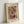 May include: Abstract watercolor artwork in light wood frame leaning against white wall. Painting features swirling organic shapes in red, green, blue, and orange with flowing black lines and geometric elements. Frame has cream matting with artist name and library text below artwork. Soft natural lighting creates gentle shadows on textured white surface.