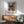 May include: Framed Van Gogh-inspired poster showing Mediterranean village with swirling night sky, yellow buildings, and cypress trees, displayed above mid-century modern wooden credenza with ceramic lamp, next to sage green armchair with walnut frame in sunlit room with white walls