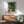 May include: Framed Van Gogh-style poster showing swirling cypress trees in rich greens and blues, displayed above a mid-century modern wooden credenza with white ceramic lamp. Light green upholstered armchair with wooden frame sits nearby on hardwood floors. Dramatic sunlight creates angular shadows across white wall in minimalist living room setting.