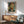 May include: Van Gogh self-portrait poster in wooden frame hanging above mid-century modern credenza with white table lamp. Teal upholstered armchair with wooden arms sits nearby. Warm sunlight creates geometric shadows on white wall. Post-impressionist artwork shows artist with reddish beard and swirling brushstroke background in blues and greens.