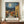 May include: Framed art print showing a Mediterranean village scene with yellow stone buildings, terra cotta roofs, and a church spire under a swirling blue night sky with bright moon. The painting uses thick brushstrokes in Van Gogh's distinctive style. Text below reads "Vincent Van Gogh" with French caption. The brown-framed poster sits on a wooden console table beside a white ceramic vase and books against cream paneled walls.