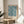 May include: Blue and cream botanical print featuring William Morris's marigold pattern displayed in light wood frame on round wooden side table next to beige upholstered chair. Natural sunlight creates soft shadows across white wall. Green plant leaves visible in corner. The intricate floral design shows repeating marigolds and foliage in vintage Arts and Crafts style with exhibition text below artwork.