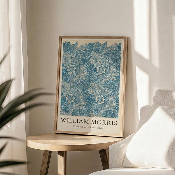 May include: Blue and cream botanical print featuring William Morris's marigold pattern displayed in light wood frame on round wooden side table next to beige upholstered chair. Natural sunlight creates soft shadows across white wall. Green plant leaves visible in corner. The intricate floral design shows repeating marigolds and foliage in vintage Arts and Crafts style with exhibition text below artwork.
