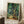 May include: A framed Van Gogh-style poster print featuring swirling cypress trees in vibrant greens, blues, and yellows with characteristic flowing brushstrokes. The artwork is displayed in a light wood frame, leaning against a white wall near a beige couch. A white coffee cup sits on a wooden side table nearby, creating a cozy living room atmosphere with natural window lighting casting soft shadows.