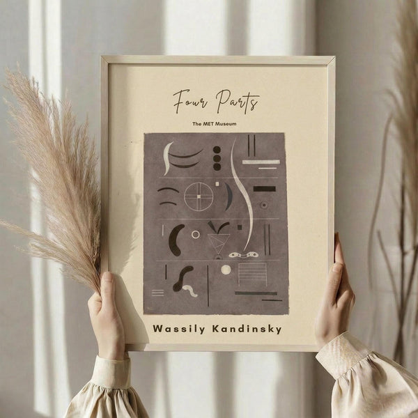 May include: Person holding framed abstract art print featuring geometric shapes and curved lines in brown and beige tones. Light wooden frame against cream background with dried pampas grass stems visible. Minimalist modern wall art in neutral earth tone palette with various abstract elements including circles, crescents, and linear forms arranged in grid-like composition.