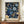May include: Framed art poster featuring intricate blue and cream floral pattern with ornate blooms and scrolling foliage on dark background, displayed on wooden sideboard next to white ceramic vase and stacked books in elegant room with cream walls and white molding