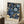 May include: Framed poster with intricate blue floral pattern featuring ornate blooms and flowing foliage on dark navy background with cream text at bottom, leaning against white wall corner next to wooden side table with white teacup and saucer, positioned on light wooden floor beside beige textured throw blanket