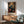 May include: Framed botanical art print featuring a blue bird perched among vibrant orange and red flowers with dark green foliage, displayed above a wooden sideboard with white ceramic lamp, next to a mint green mid-century modern armchair with wooden arms in a bright room with natural sunlight casting shadows on white walls.