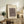 May include: Framed beige poster with abstract geometric shapes in brown tones displayed on rustic wooden sideboard next to fern plant, ceramic vase, and stacked books in warm neutral living room setting with cream upholstered chair visible in foreground.