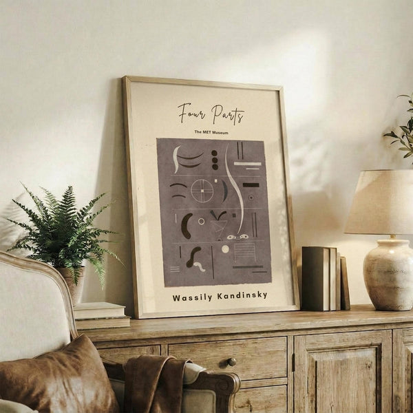 May include: Framed beige poster with abstract geometric shapes in brown tones displayed on rustic wooden sideboard next to fern plant, ceramic vase, and stacked books in warm neutral living room setting with cream upholstered chair visible in foreground.