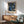 May include: Framed botanical poster with intricate blue and cream floral pattern on dark background, displayed above wooden sideboard with white ceramic lamp. Mid-century modern sage green armchair with wooden arms sits nearby on light wooden floors. Natural sunlight creates geometric shadows on white wall, creating warm, contemporary living room setting.