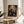 May include: Framed poster featuring a blue bird perched among ornate botanical patterns with red and coral flowers on a dark teal background, displayed on a round wooden side table next to a beige upholstered chair. Soft natural lighting creates shadows on white walls, with a plant visible in the corner of the room.