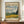 May include: Van Gogh museum poster featuring Wheat Field with Cypresses painting in brown frame, displayed on wooden credenza beside white ceramic vase and stacked books. Poster shows swirling blue-green sky above golden wheat field with dark cypress trees. Cream background with black text including museum attribution and painting title.