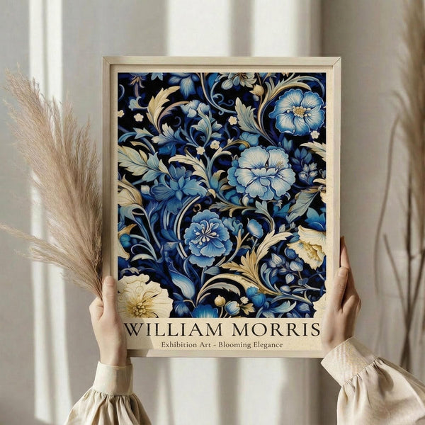 May include: Person holding framed poster with ornate blue and cream floral pattern in decorative style, featuring blooming flowers and swirling botanical motifs on dark background. Light wooden frame surrounds artwork with William Morris exhibition text below. Dried pampas grass visible beside figure wearing cream ruffled sleeves against neutral curtained background.